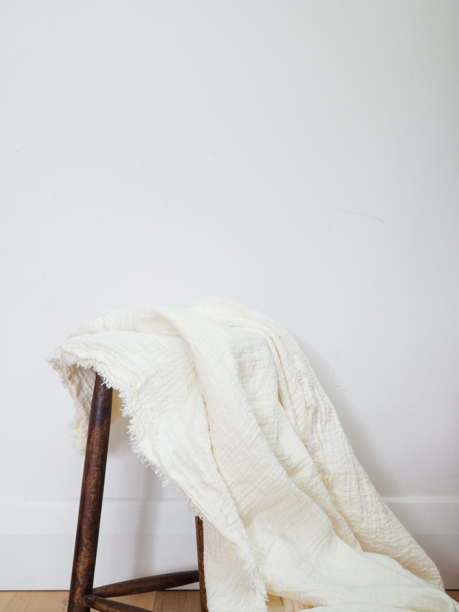 Heavyweight linen blanket in white flowing from a rustic stool to show its large length and fringed detailing.